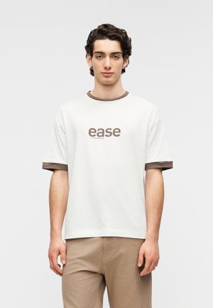 Hvid t-shirt i bomuld med brun ribkant på kraven og ærmekanter; har teksten "ease" i brun på forsiden. Bærtes med lysebrune bukser.