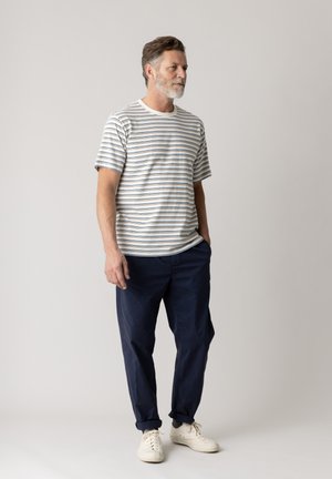 Mann mit grauem Haar und Bart, der ein gestreiftes kurzärmeliges Hemd, marineblaue Hose und weiße Sneakers trägt, steht mit den Händen in den Taschen.