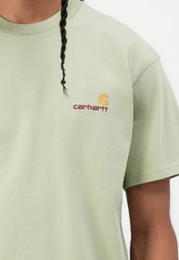 Light green cotton t-shirt with short sleeves, featuring a curved logo in yellow and "carhartt" in maroon embroidered on the chest.