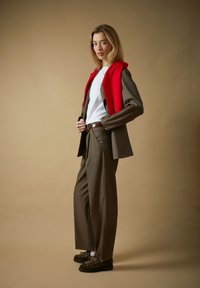 Ung kvinne i brun dress og hvit skjorte, med rød genser kastet over skuldrene, stående foran en ensfarget beige bakgrunn.