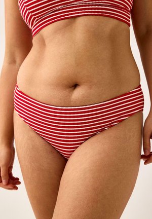 Kvinde iført rød- og hvidstribet bikiniunderdel og -top, der viser mellemgulvet og hænder mod en neutral baggrund.