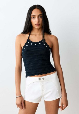 Femme aux longs cheveux foncés portant un haut dos nu noir avec des détails œillets et un short blanc taille haute sur un fond uni.