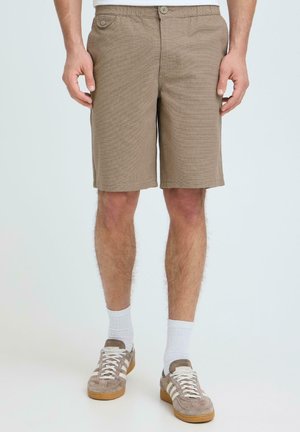 Beige afslappede shorts båret af en stående mand med hvide crew-sokker og beige ruskindssneakers med gummisåler.
