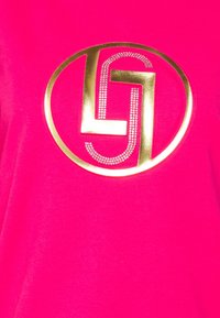 Horkorůžové tričko s velkým zlatým kruhovým logem se stylizovaným "G" a akcenty z rhinestone. Hladká textura, krátké rukávy.