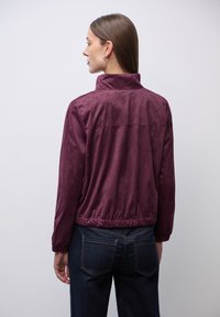 Burgundská semišová bunda s vysokým límcem, elastickým lemem a dlouhými rukávy. Na zadní straně se nachází horizontální šev.