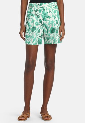 Kvinde iført højttaljede hvide shorts med grønt blomstertryk, kombineret med brune flade sandaler med stropper, stående mod en ensfarvet baggrund.