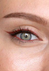 Närbild av ett grönt öga med långa ögonfransar, med röd eyeliner. Huden har en jämn textur och subtila höjdpunkter.