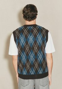 Schwarze und blaue Argyle-Strickweste mit weißen Akzenten, geripptem Kragen und Saum, über einem weißen T-Shirt, von hinten präsentiert.