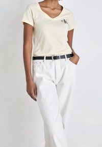 Camiseta amarilla clara de cuello en V con mangas cortas, que presenta un pequeño logo negro, combinada con pantalones blancos de cintura alta y un cinturón negro.