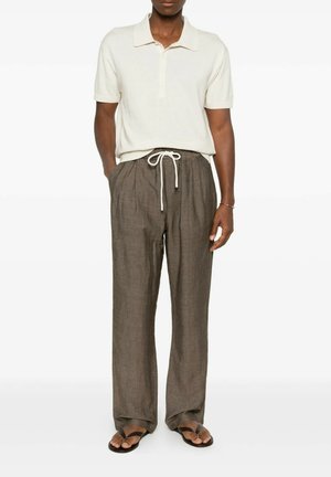 Man draagt een wit poloshirt met korte mouwen, losse bruine broek met trekkoord en zwarte slippers, staand met één hand in de zak.