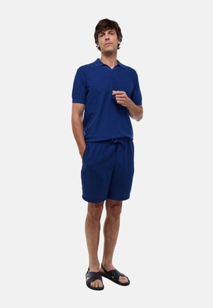 Tenue masculine composée d'un polo à manches courtes bleu uni et d'un short à cordon assorti, accompagnés de sandales noires. Tissu texturé.