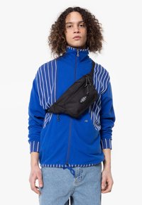 Marsupio nero in tessuto indossato in diagonale su una giacca blu con zip e strisce bianche, con spallacci regolabili e un piccolo logo.