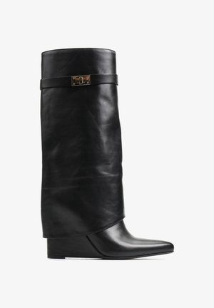 Czarne skórzane botki na koturnie z luźniejszą cholewą, z płaskim paskiem i metalowym akcentem. Spiczasty nosek z gładką powierzchnią.