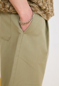 Light olive green pants with side pockets, featuring a smooth texture. A silver chain bracelet adorns the wrist in the pocket area.