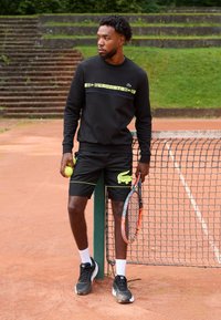 Mann i svart sportbekledning som holder tennisballer og racket, lener seg mot nettet ved et leirtennisbane med trapper og gress i bakgrunnen.