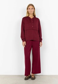 Hoodie bordeaux avec poche avant et boutons, associé à un pantalon large assorti. Le tissu semble doux et texturé. Ballerines marron avec des clous.