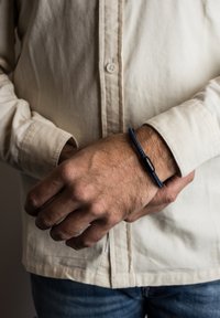 Bracciale intrecciato blu con un accento di perla nera indossato su un polso, mani appoggiate su una camicia leggera a bottoni.