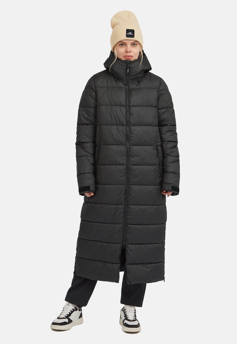 Sort dunjakke med et langt, lige design, der har en høj krave og lynlåslommer. Kombineret med en beige beanie og sneakers.