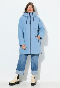 Ljusblå vattentät parkas med huva, framdragkedja, två sidofickor och svarta snoddar. Bärs över avkortade denimjeans och stövlar.
