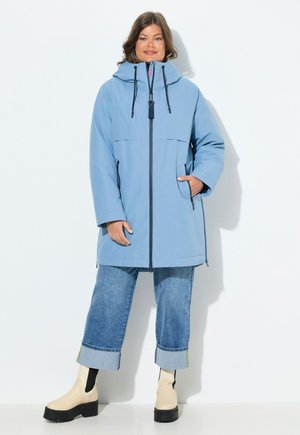 Parka imperméable bleu clair avec capuche, fermeture éclair à l'avant, deux poches latérales et cordons de serrage noirs. Porté par-dessus un jean en denim raccourci et des bottes.