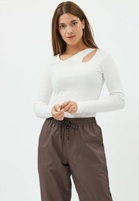 Hvid ribbet langærmet top med udskæringer ved skulderen, parret med løse brune bukser med snørelukning. Glat tekstur, tætsiddende design.