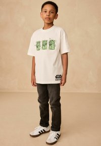 Tricou alb din bumbac cu trei desene grafice verzi și textul "Star Wars Mandalorian" în negru. Asociat cu blugi închisi la culoare și adidași albi.