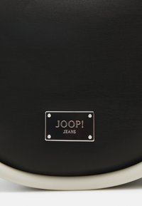 Bolso de cuero sintético negro que presenta una placa plateada con "JOOP! JEANS" en texto en relieve. Textura suave y forma redondeada con acentos ligeros.