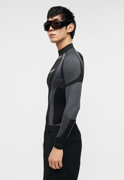 Μαύρο αθλητικό τοπ μακρυμάνικο με γκρι υφασμάτινες ρίγες, εφαρμοστή σχεδίαση και ψηλό γιακά. Φοριέται με μεγάλα σκουρόχρωμα γυαλιά ηλίου.