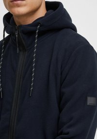 Sudadera con capucha de forro polar azul marino con cremallera completa, cordones ajustables con acentos amarillos y un parche de logo en el lateral. Tejido suave y texturizado.