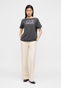 Szara bawełniana koszulka z logo "EA7 Emporio Armani", zestawiona z kremowymi, szerokimi spodniami z bocznymi detalami oraz czarnymi szpilkami.