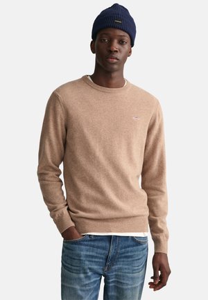 Beige gebreide trui met een ronde hals, lange mouwen en geribbelde manchetten. Draagt over een wit shirt. Gecombineerd met blauwe denim jeans.