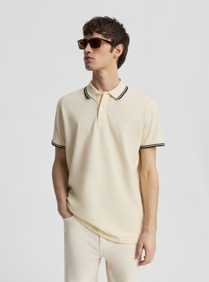 Młody mężczyzna ubrany w beżową koszulkę polo z czarnym paskowanym kołnierzykiem i rękawami, beżowe spodnie i brązowe okulary przeciwsłoneczne, stoi z jedną ręką w kieszeni.