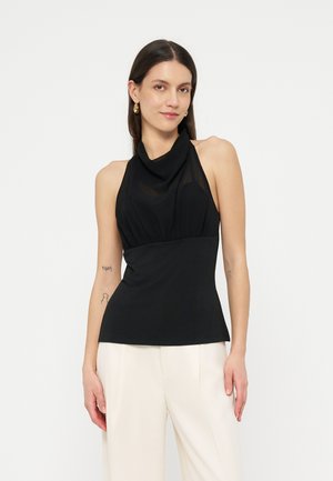 Femme portant un haut noir sans manches à col halter et un pantalon blanc, se tenant devant un fond clair uni, avec des boucles d'oreilles créoles dorées.