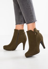 Bottes de cheville en daim vert olive avec un bout pointu, un talon de 10 cm de hauteur, une fermeture éclair sur le côté et un accent de boucle à la cheville. Surface texturée.