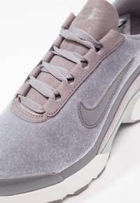 Baskets en daim gris clair avec lacets, panneau texturé gris et semelle en caoutchouc blanc. Présente un logo visible sur la languette et sur le côté.