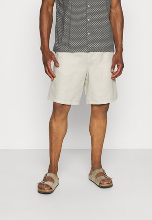 Homme portant un short beige, une chemise à manches courtes boutonnée grise à motifs, et des sandales à boucle marron clair, debout avec les mains détendues le long du corps.