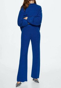 Blauwe gebreide trui en bijpassende wijde broek. De stof is gestructureerd, met een losse pasvorm en een hoge hals. Sprankelende schoenen maken de outfit compleet.