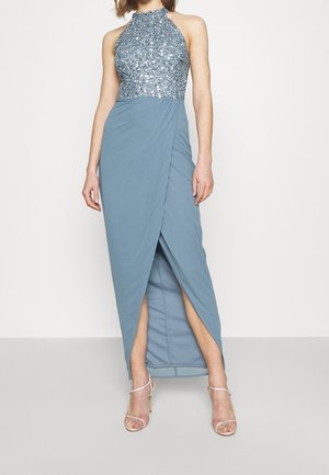 Kobieta w niebieskiej, bezrękawowej sukni z cekinowym topem na szyi i rozcięciem z przodu, w połączeniu z sandałkami na wysokim obcasie z paskami.