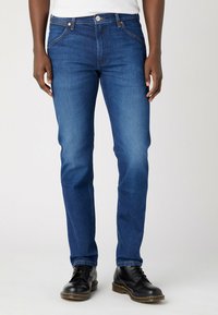 Jeans denim w ciemnoniebieskim odcieniu z prostą nogawką, posiadające pięć kieszeni, miedziane przeszycia i zapięcie na guzik. Widać czarne buty.