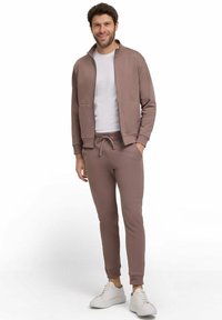 Brauner Sweatshirt mit Reißverschluss vorne, zwei seitlichen Taschen, kombiniert mit passenden schmal geschnittenen Jogginghosen und weißen Sneakern. Glatter, lässiger Stoff.