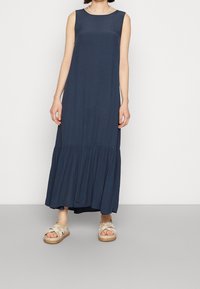 Nainen, jolla on yllään säädyttömät tummansiniset maksimekot, joissa on kerätty helma, yhdistettyinä beigeihin ristikkäisiin sandaaleihin.