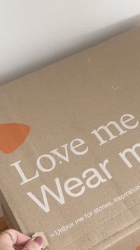 Bruine kartonnen doos met witte tekst, voorzien van een oranje accent. De tekst bevat "Hou van me, Draag me," en "Open mij voor verhalen, inspiratie."