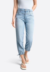 Vaqueros azul claro de talle alto con un corte relajado, dobladillo deshilachado y lazos ajustables en los tobillos, combinados con chanclas blancas.