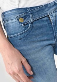 Mano apoyada junto a la cadera, vistiendo jeans de mezclilla azul con un cinturón ancho abrochado con un botón de latón, combinados con una camiseta blanca acanalada.