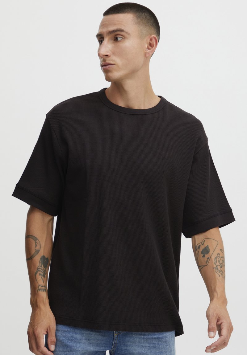 Sort kortærmet t-shirt i et struktureret stof med rund halsudskæring og rullede ærmer, parret med blå jeans, der viser en tatoveret arm.