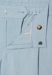 Pantaloni azzurri chiari con design plissettato, vita elastica e chiusura a bottone. Cuciture visibili e texture liscia.
