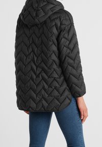 Giacca a vento nera con cappuccio, caratterizzata da un motivo a zig zag e un orlo arrotondato. Indossata sopra dei jeans blu.