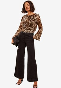 Blouse à imprimé léopard avec de longues manches évasées, associée à un jean noir à taille haute et jambes larges. Sac à main noir, sandales à talons ouvertes.