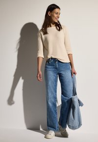 Beige strukturierter Pullover mit 3/4-Ärmeln, kombiniert mit hellblauen, weit geschnittenen Jeans. Dazu wird eine helle Denim-Tasche gehalten; weiße Sneakers runden das Outfit ab.