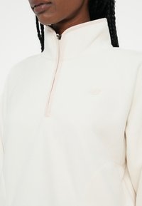 Light cream fleece pullover with a high collar and half-zip design. Features subtle tonal logo and soft texture, suitable for layering.